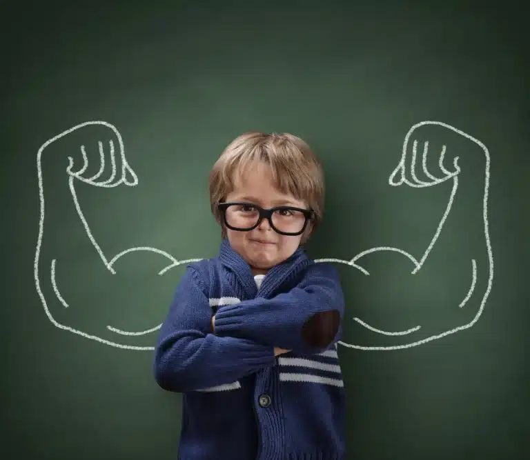 A young boy in glasses and a blue sweater stands in front of a chalkboard with muscular arms drawn behind him.